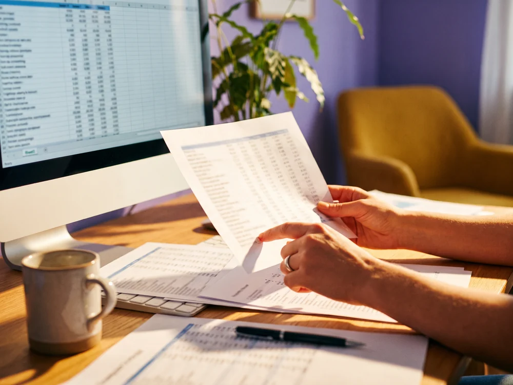 Hands holding a printed financial report beside a monitor of matching data, checking that the two sets of figures reconcile