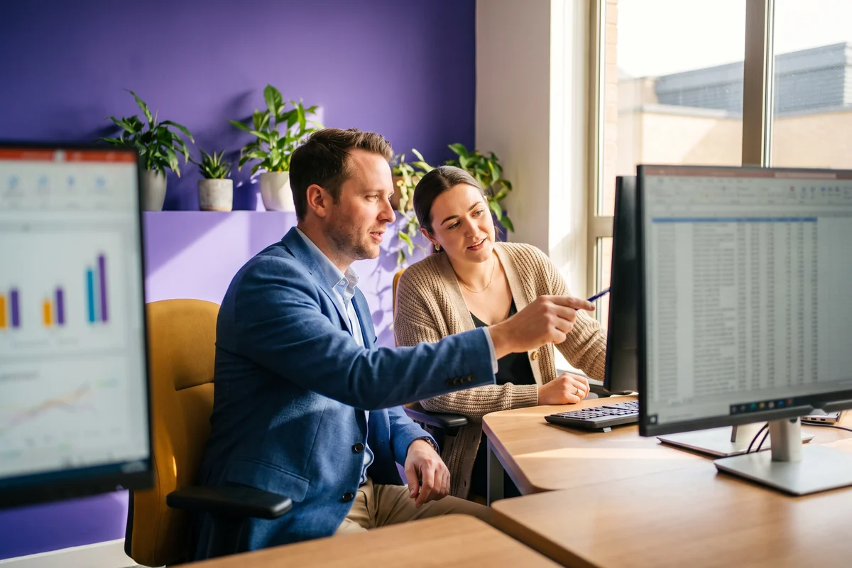 A Power BI consultant pointing at the underlying data on one screen while a dashboard of charts sits on the other, showing where a number really comes from
